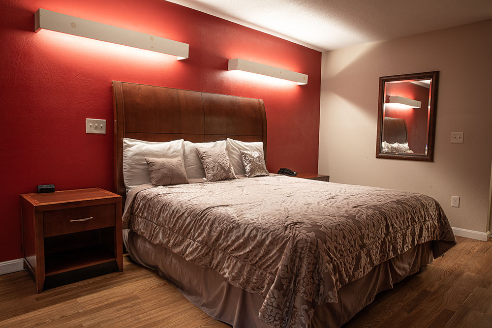 A single bed with beautiful wooden headboard against a red wall. Two over head lights, rectangular in shape, above the bed. There's a bedside table as well