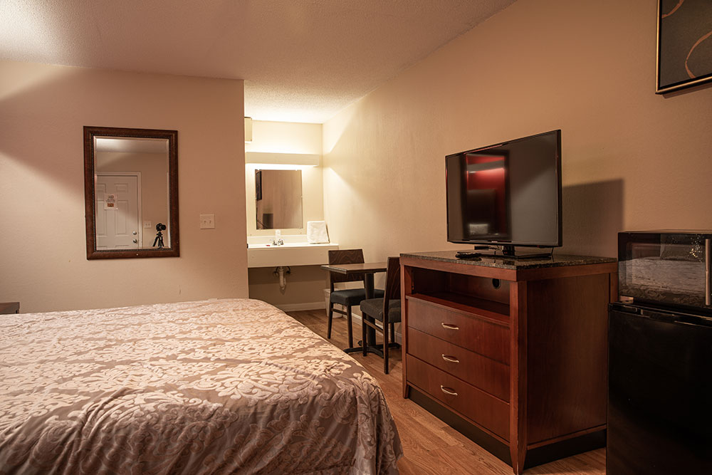 The same room as above but showing the foot of the bed, the opening to the bath area, a dresser, flat screen tv, table and chairs